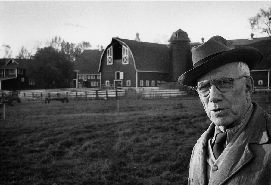 Man in overcoat and fedora. Barn yard in background. Black and white photo.