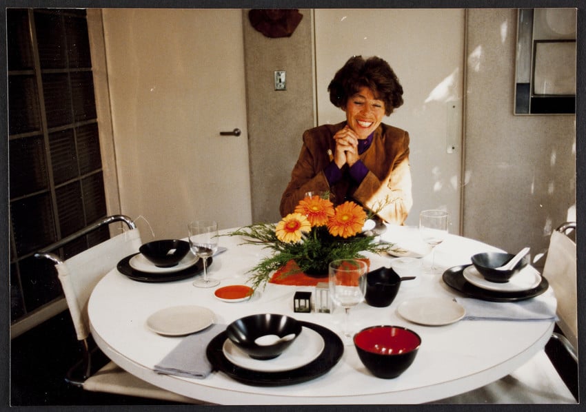 Ati Gropius sitting at the dining table at the Gropius House, set with dishes and a bouquet of Gerber daisies..