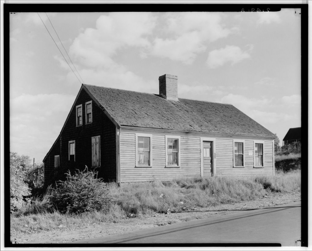 Black-and-white photo of a wooden house with a gabled roof and chimney, adjacent to a road.