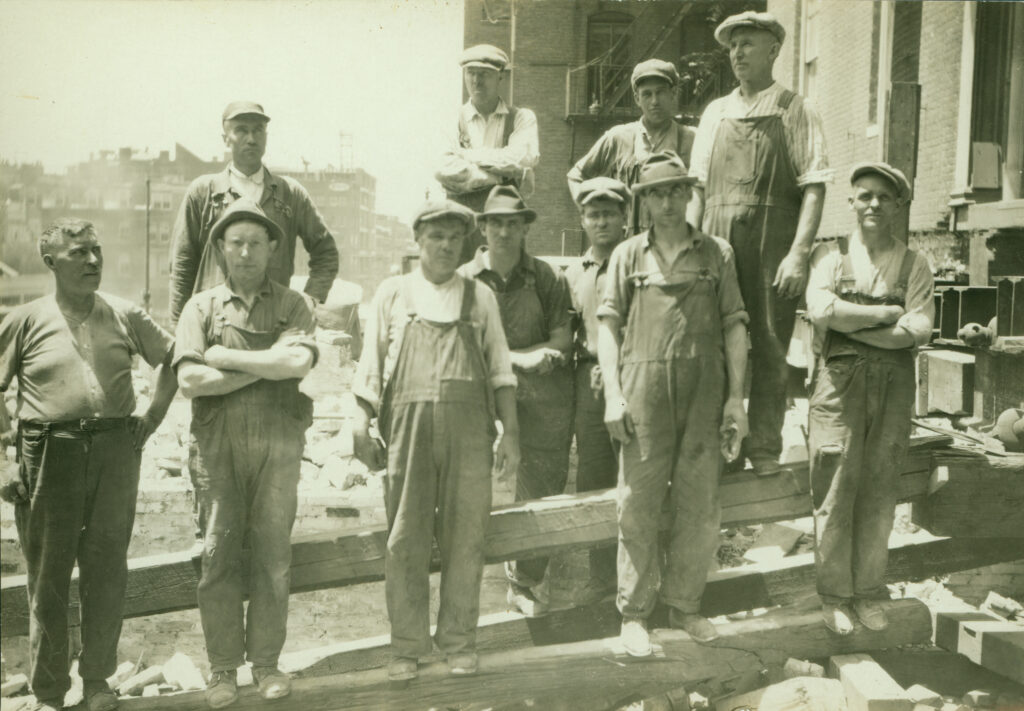A group of eleven men in work overalls and caps at a construction site.