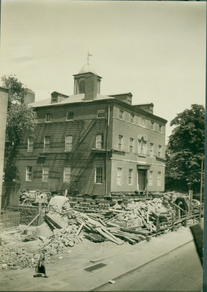 Construction site around a three-story brick building with fire escape stairs and a cupola.